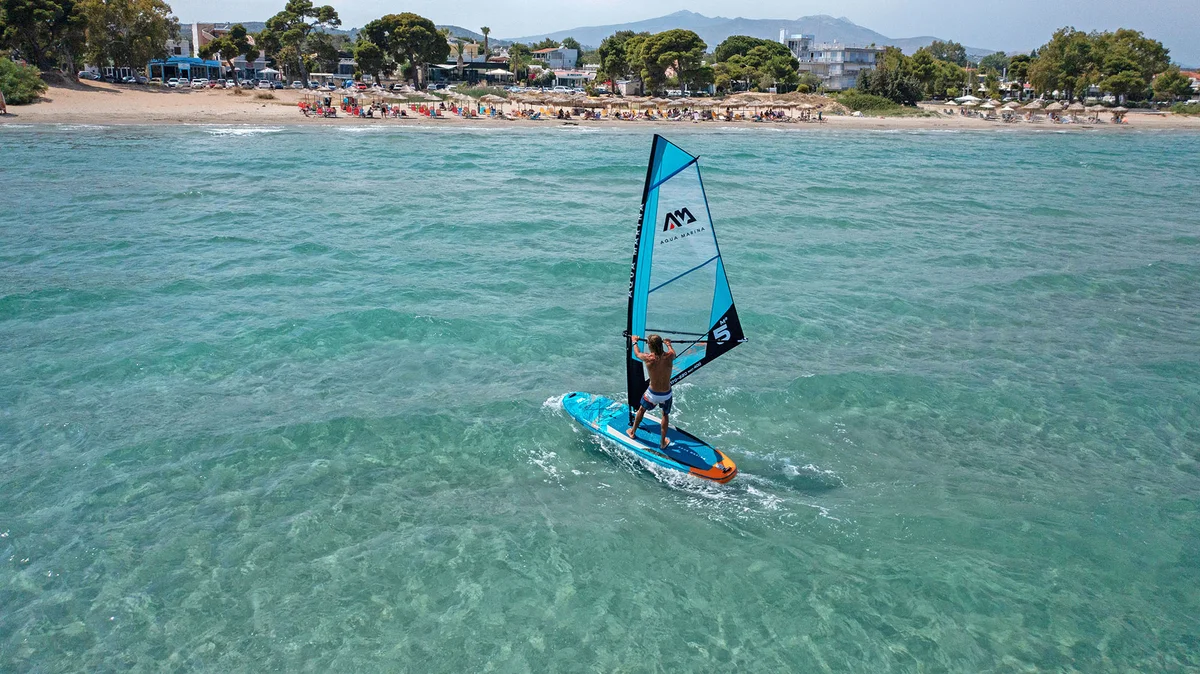 Mężczyzna pływający na pompowanej desce WindSUP Aqua Marina Blade z żaglem przy piaszczystej plaży na turkusowym morzu