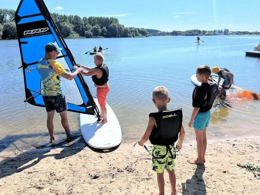 Szkolenie windsurfingowe dla dzieci na jeziorze – instruktor pomaga chłopcu na desce z małym pędnikiem, obok stoją dzieci w kamizelkach asekuracyjnych na plaży